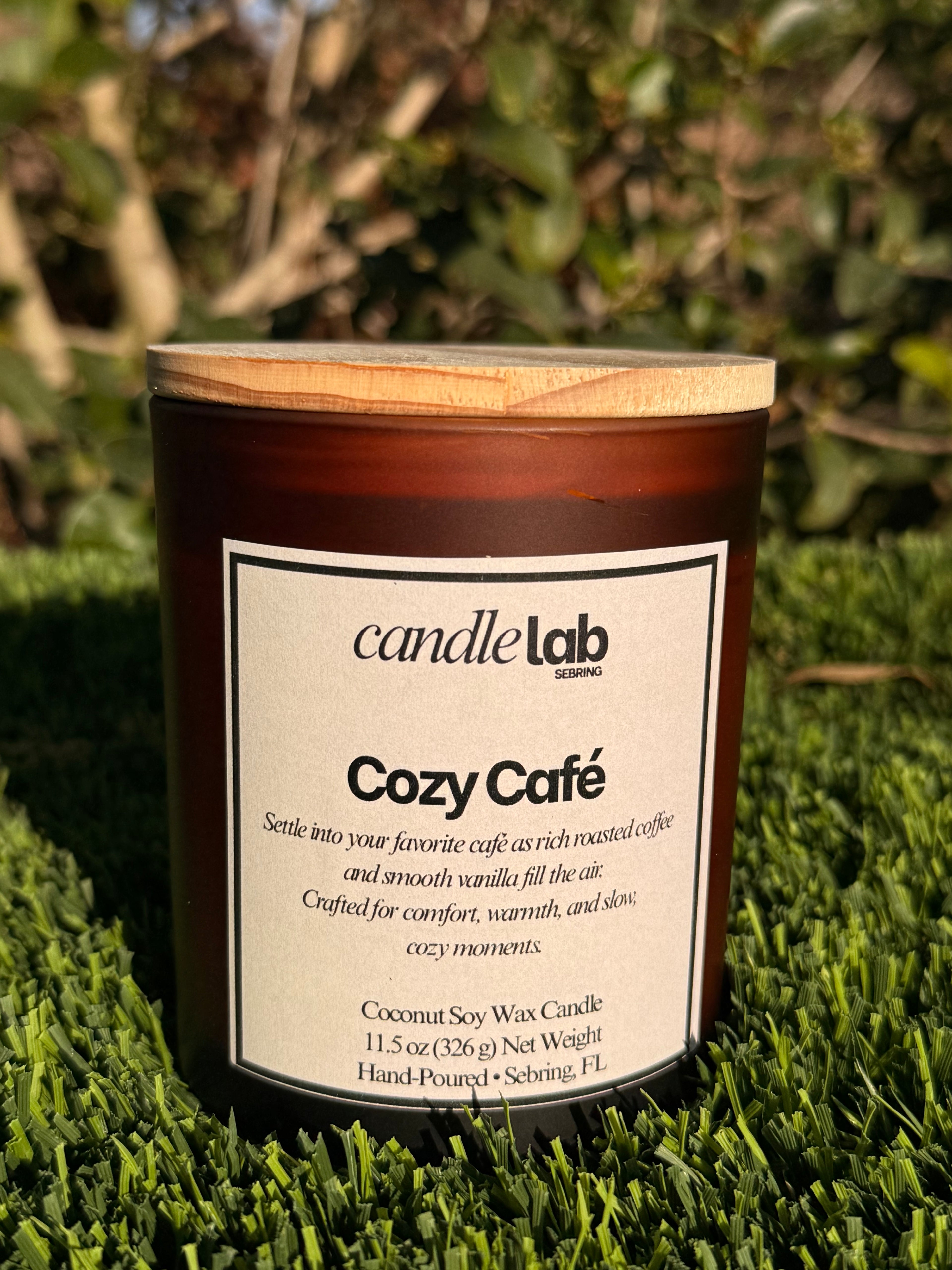 An amber glass candle with a label that says "Cozy Cafe" along a short blurb reading "Settle into your favorite cafe as rich roasted coffee and smooth vanilla fill the air. Crafted for comfort, warmth, and silent, cozy moments."