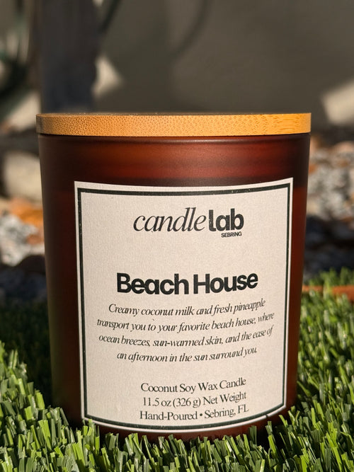 An amber glass candle with a label that says "Beach House" along with a short blurb reading "Creamy coconut milk and fresh pineapple transport you to your favorite beach house, where ocean breezes, sun-warmed skin, and the ease of an afternoon in the Sun surround you."
