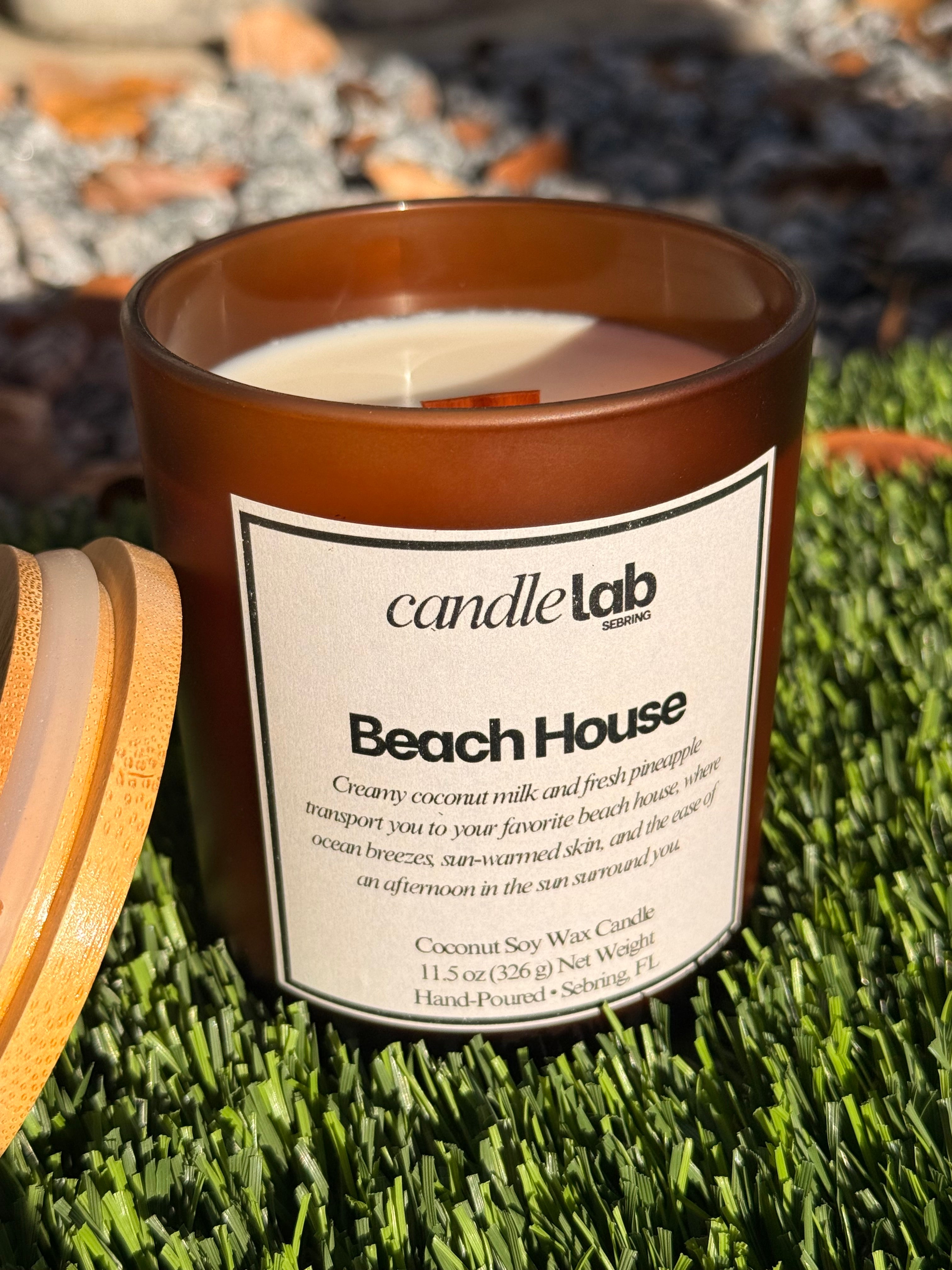 An amber glass candle with a label that says "Beach House" along with a short blurb reading "Creamy coconut milk and fresh pineapple transport you to your favorite beach house, where ocean breezes, sun-warmed skin, and the ease of an afternoon in the Sun surround you."