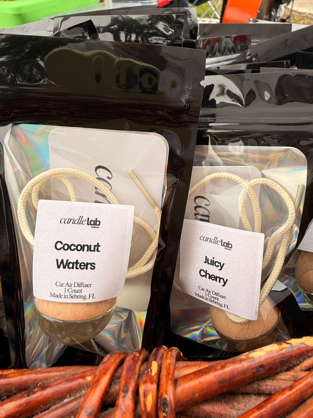 Picture of two small bags with a car diffuser inside. One is labeled "Coconut Waters" on the front and the other reads "Juicy Cherry". Both read with the information: "Car Air Diffuser. 1 Count. Made in Sebring, FL"