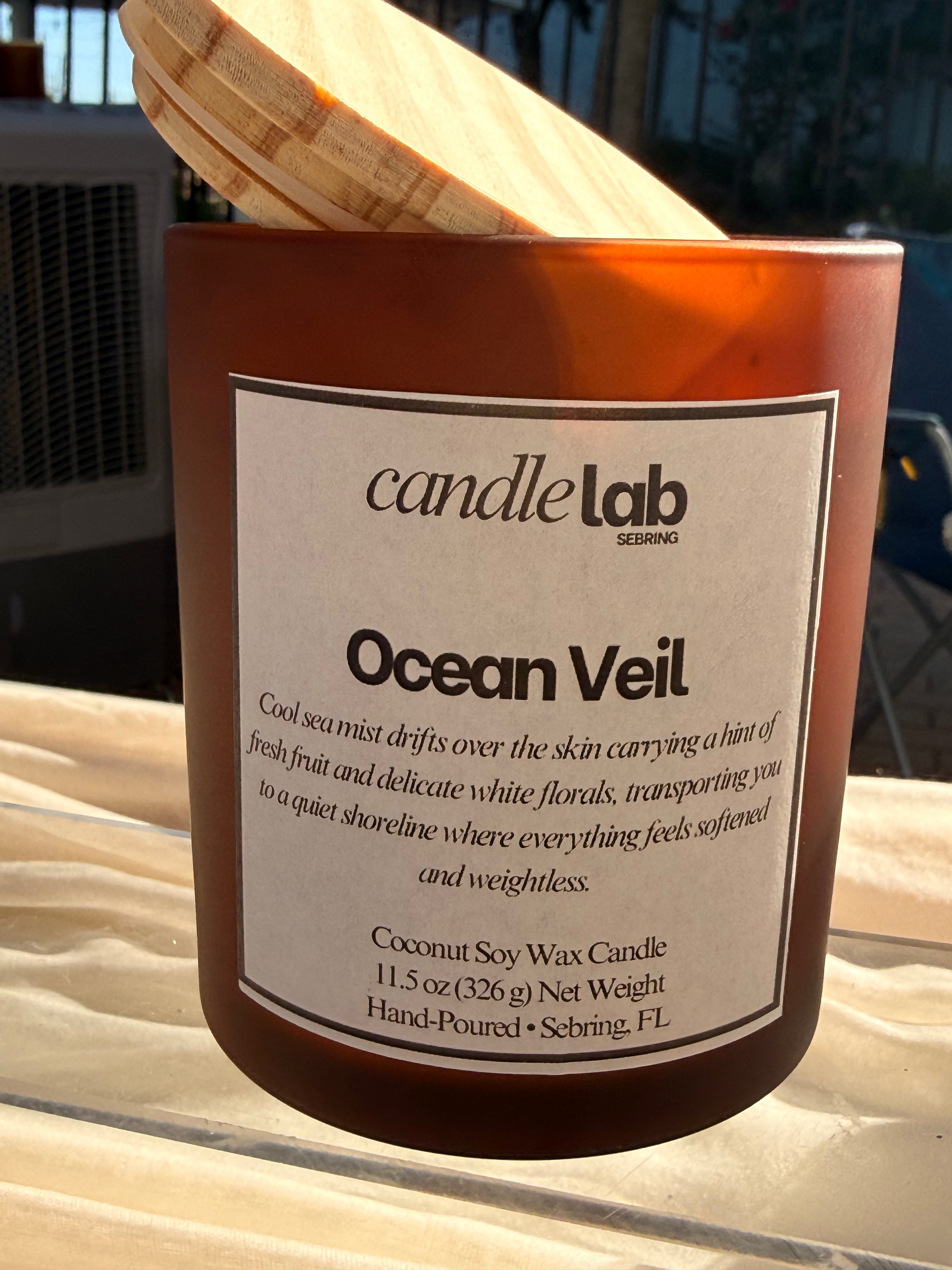 An amber glass candle with a label that says "Ocean Veil" along with a short blurb reading "Cool sea mist drifts over the skin carrying a hint of fresh fruit and delicate white florals, transporting you to a quiet shoreline where everything feels softened and weightless."