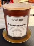 An amber glass candle with a label that says "Parisian Macaron" along with a blurb reading "Like a quiet morning in a Parisian patisserie, where the air is filled with warm almond, soft spice, and melting vanilla sweetness. This burn feels cozy, golden, and gently indulgent."