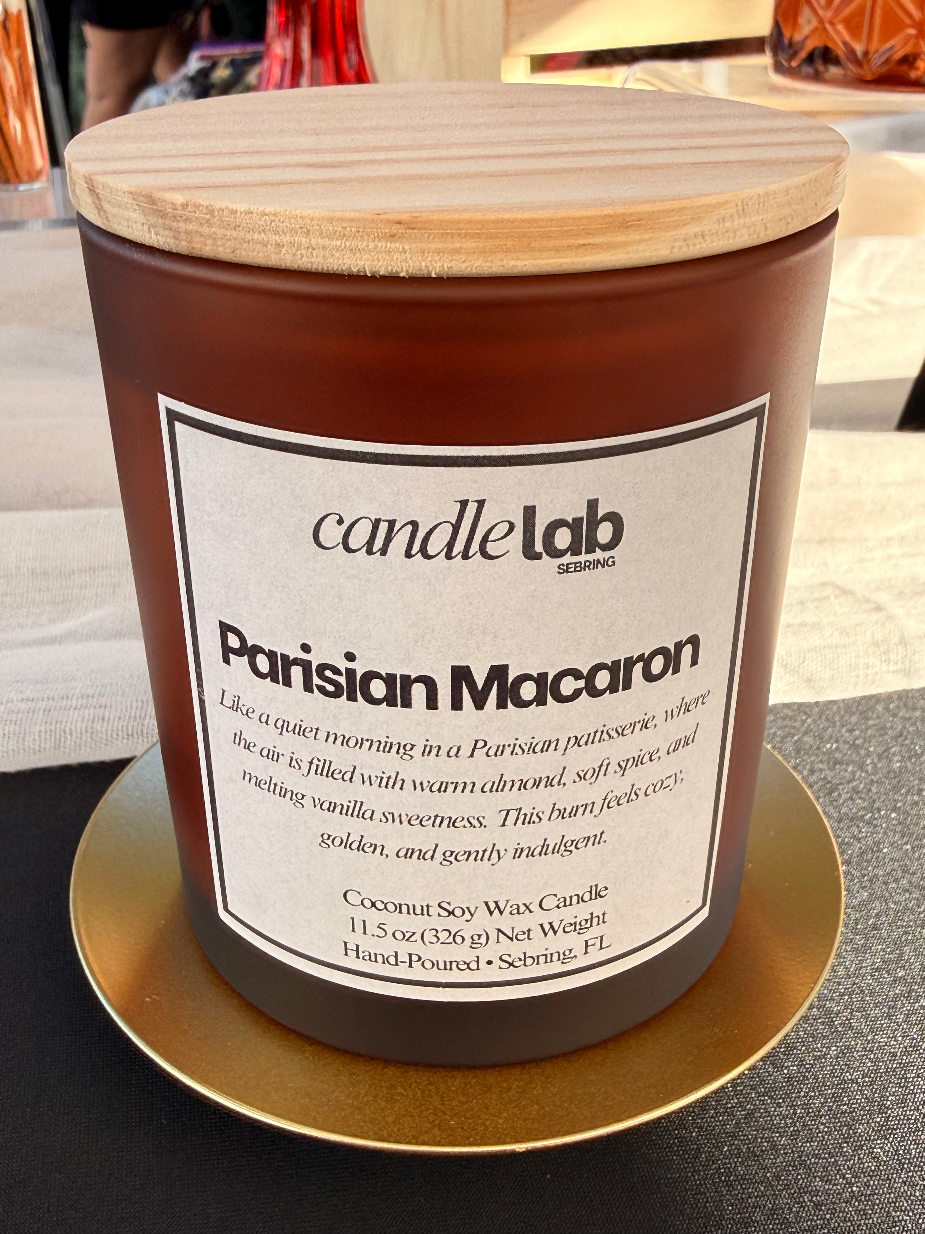 An amber glass candle with a label that says "Parisian Macaron" along with a blurb reading "Like a quiet morning in a Parisian patisserie, where the air is filled with warm almond, soft spice, and melting vanilla sweetness. This burn feels cozy, golden, and gently indulgent."