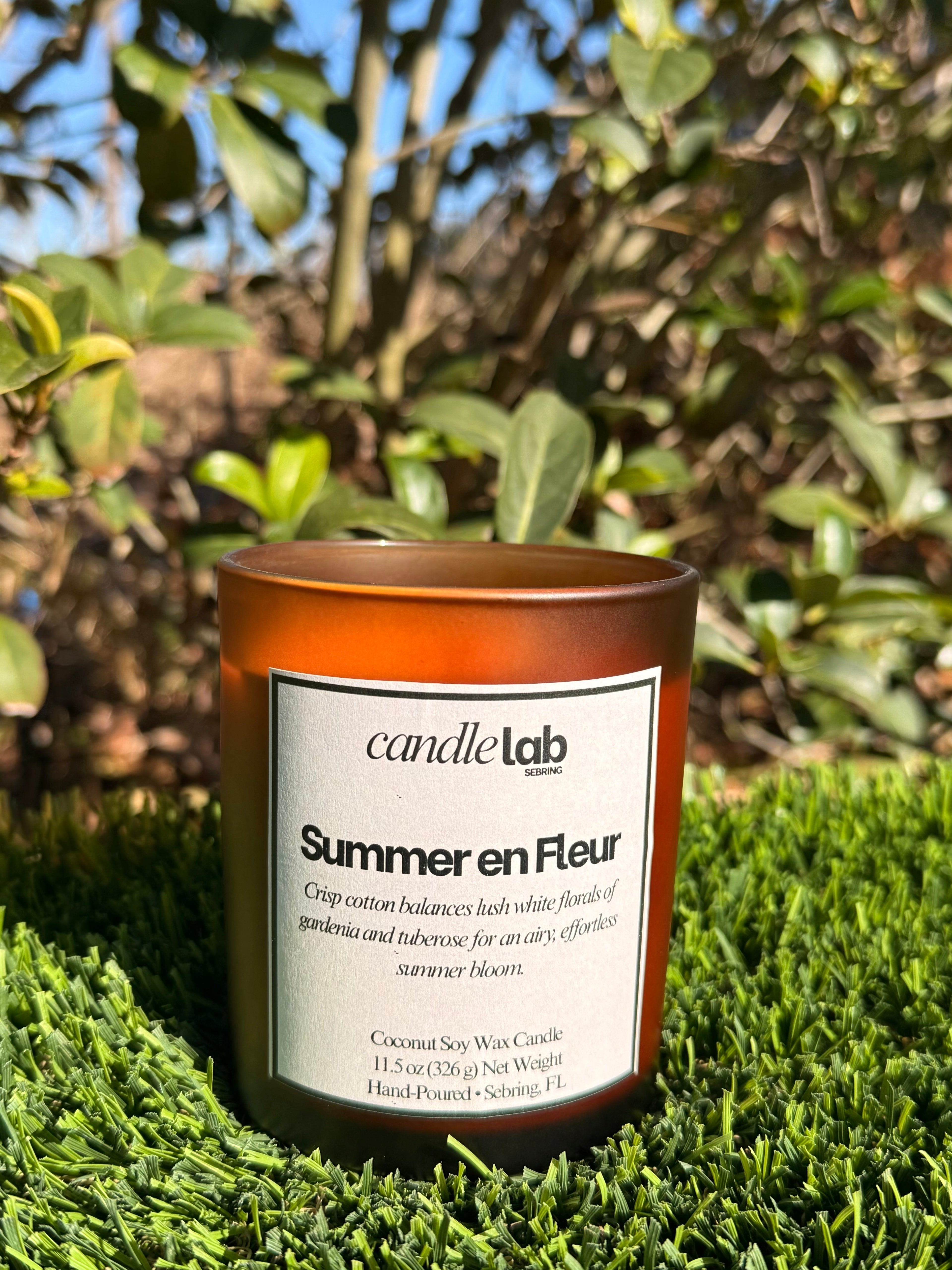 An amber glass candle with a label that says "Summer en Fleur" along with a short blurb that reads "Crisp cotton balances lush white florals of gardenia and tuberose for an airy, effortless summer bloom."
