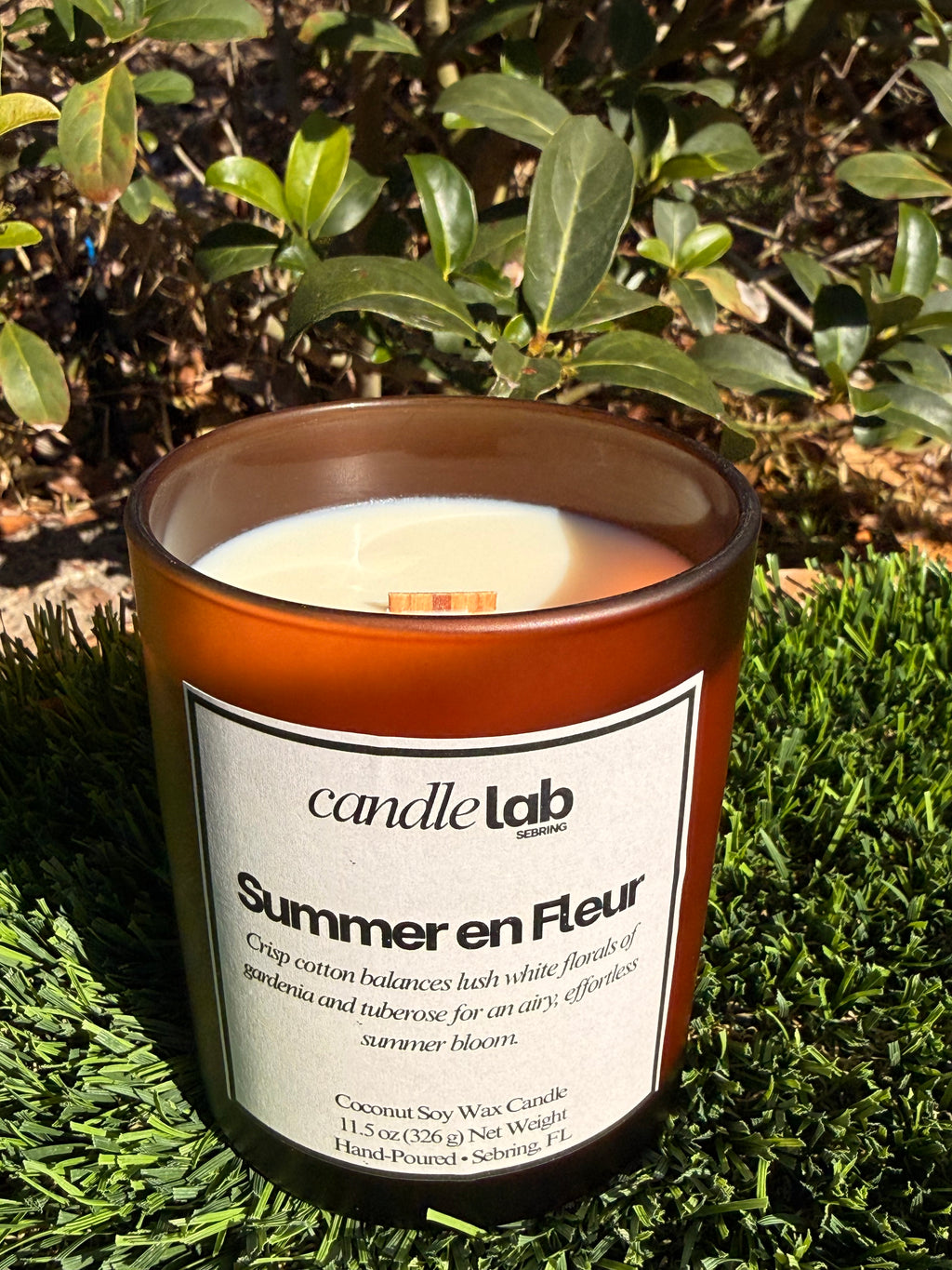 An amber glass candle with a label that says "Summer en Fleur" along with a short blurb that reads "Crisp cotton balances lush white florals of gardenia and tuberose for an airy, effortless summer bloom."
