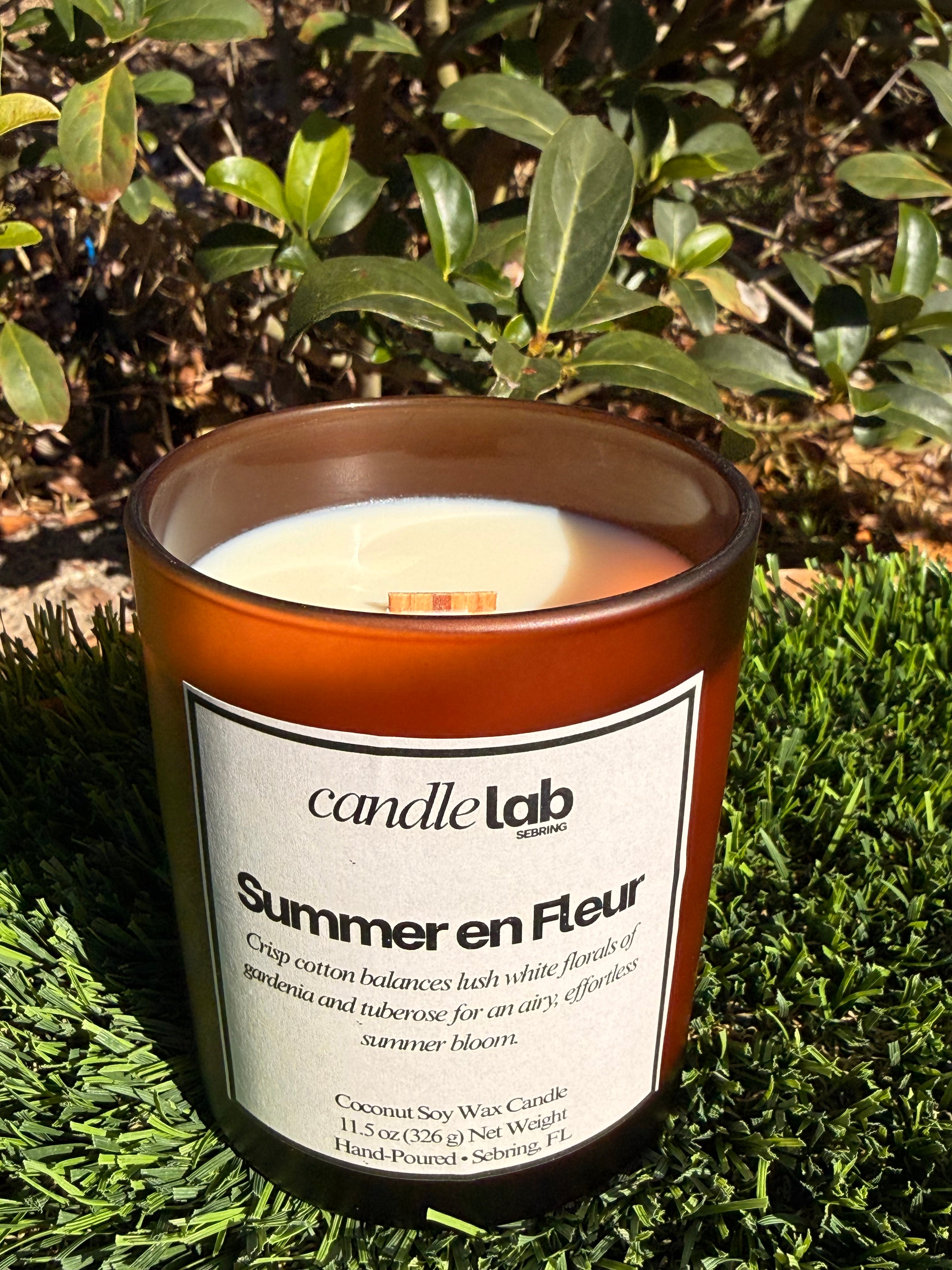 An amber glass candle with a label that says "Summer en Fleur" along with a short blurb that reads "Crisp cotton balances lush white florals of gardenia and tuberose for an airy, effortless summer bloom."