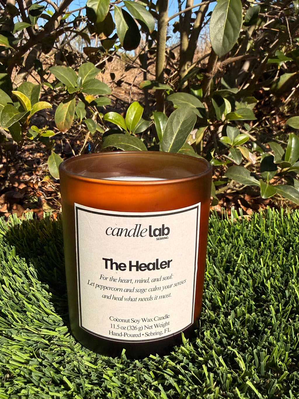 An amber glass candle with a label that says "The Healer" along with a short blurb that reads "For the heart, mind, and soul: Let peppercorn and sage calm your senses and heal what needs it most."