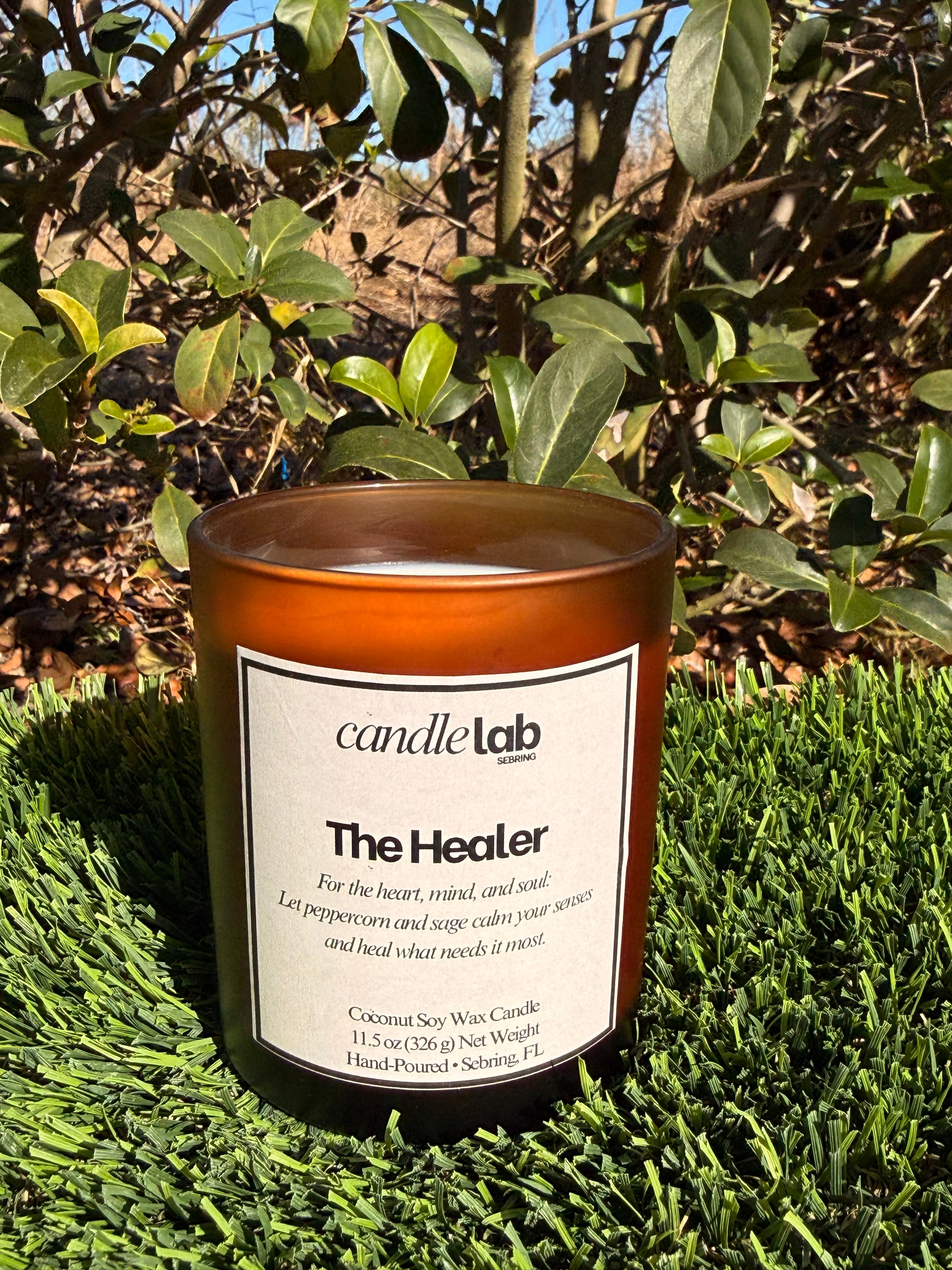 An amber glass candle with a label that says "The Healer" along with a short blurb that reads "For the heart, mind, and soul: Let peppercorn and sage calm your senses and heal what needs it most."