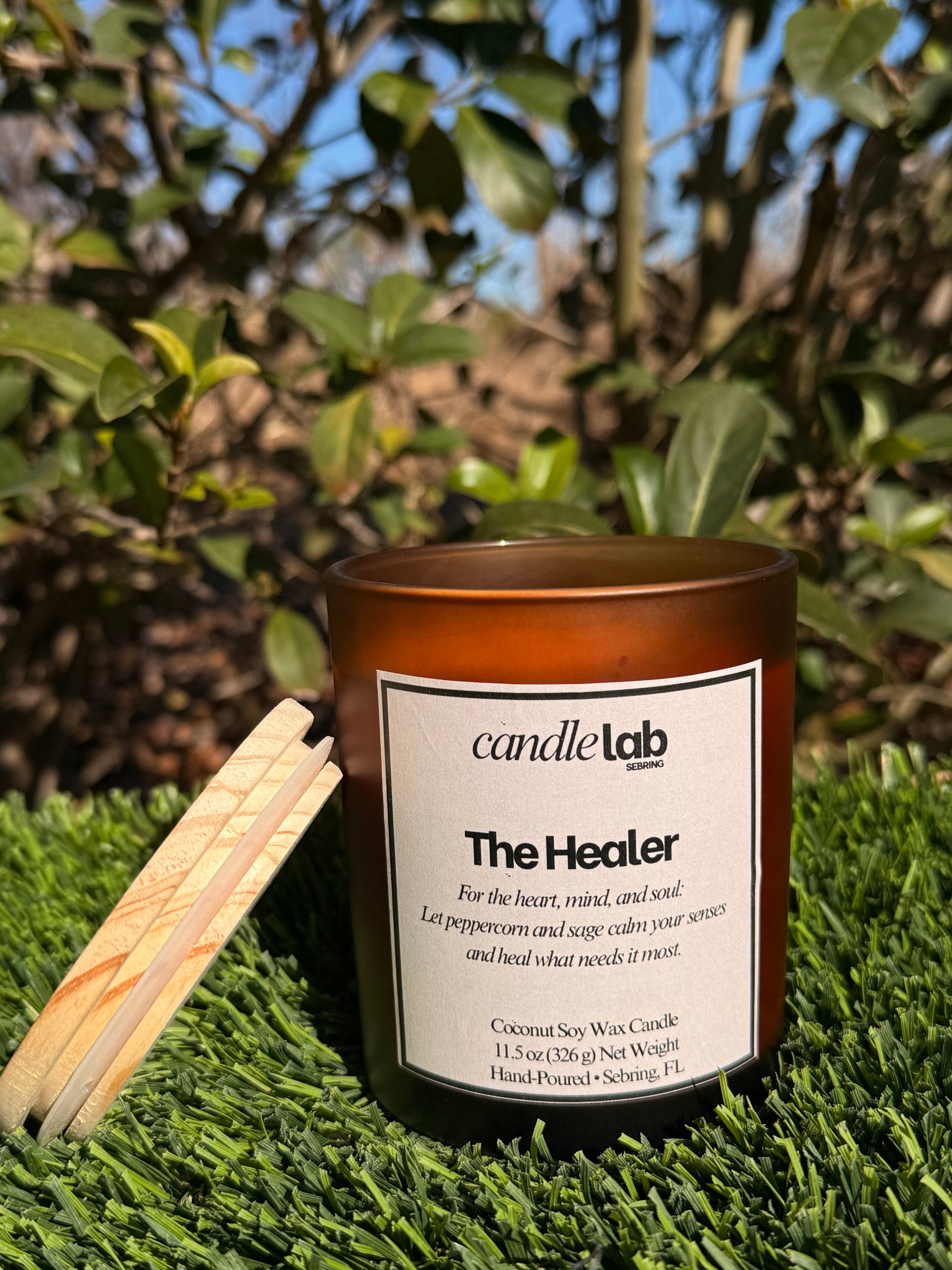 An amber glass candle with a label that says "The Healer" along with a short blurb that reads "For the heart, mind, and soul: Let peppercorn and sage calm your senses and heal what needs it most."