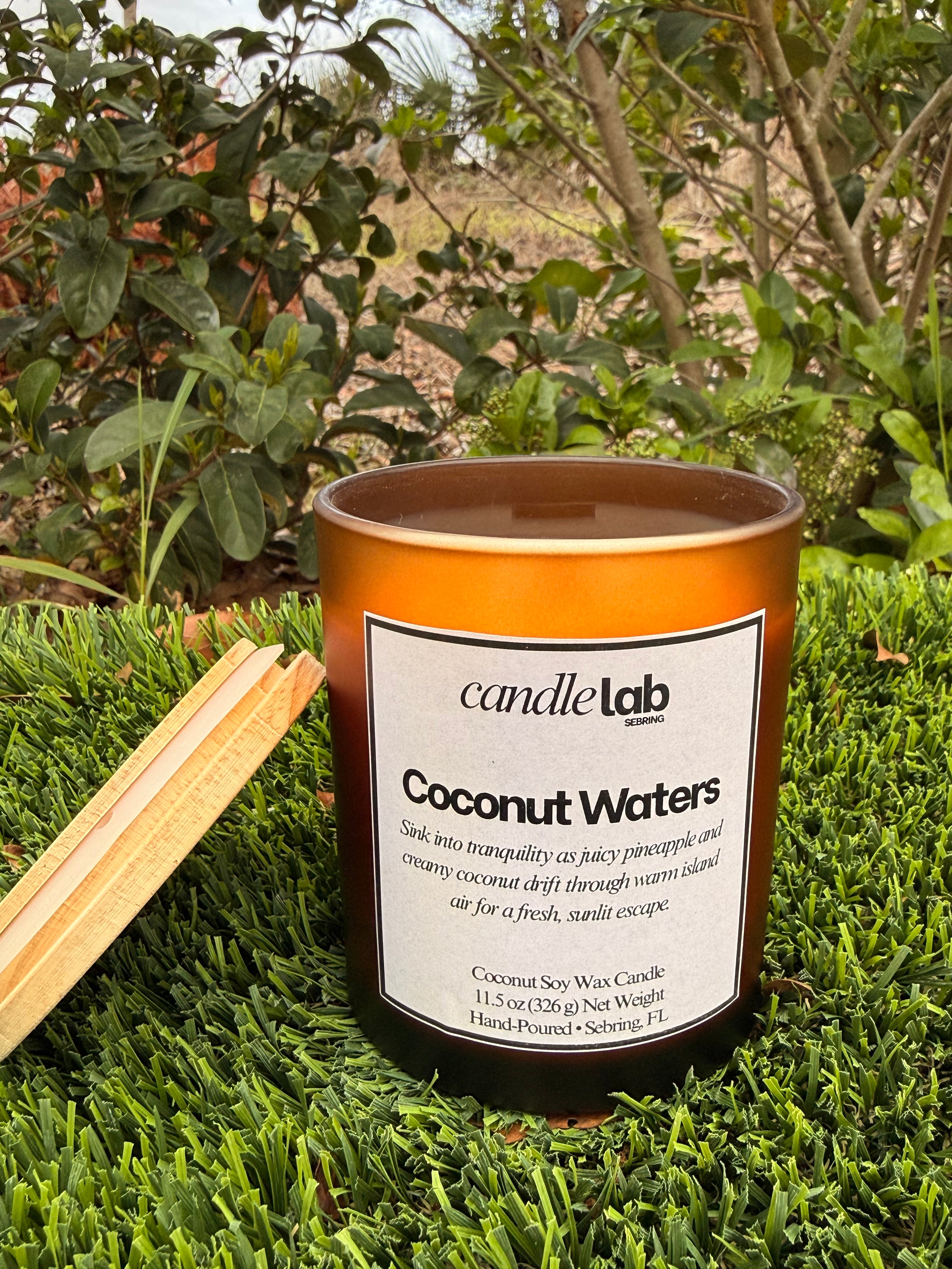 An amber glass candle with a label that says "Coconut Waters" along with a short blurb that says "Sink into tranquility as juicy pineapple and creamy coconut drift through warm island air for a fresh sunlit escape."