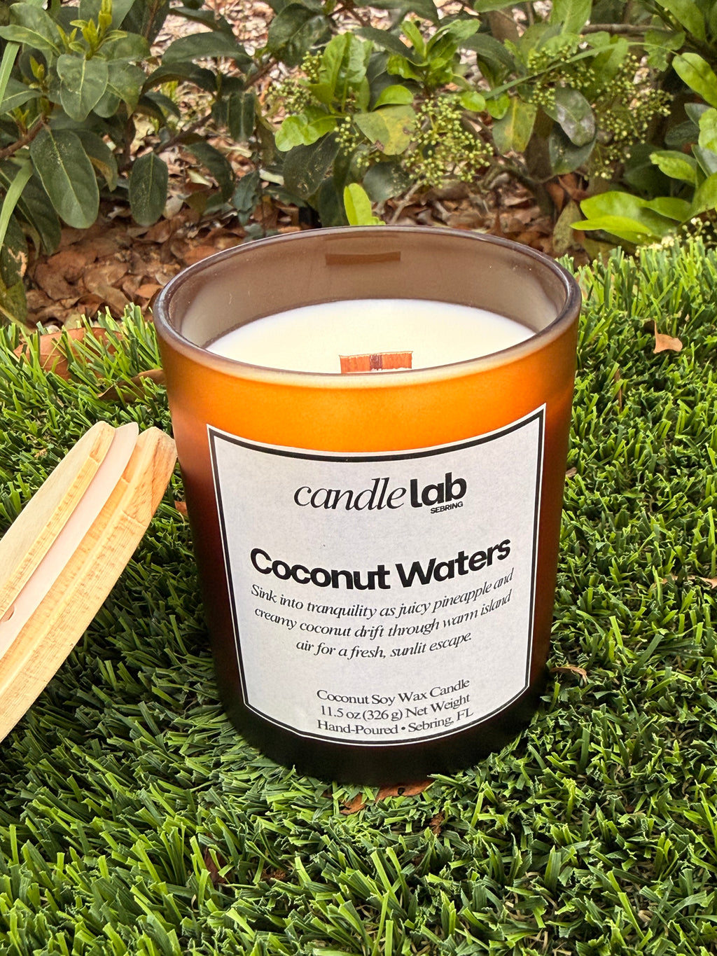 An amber glass candle with a label that says "Coconut Waters" along with a short blurb that says "Sink into tranquility as juicy pineapple and creamy coconut drift through warm island air for a fresh sunlit escape."