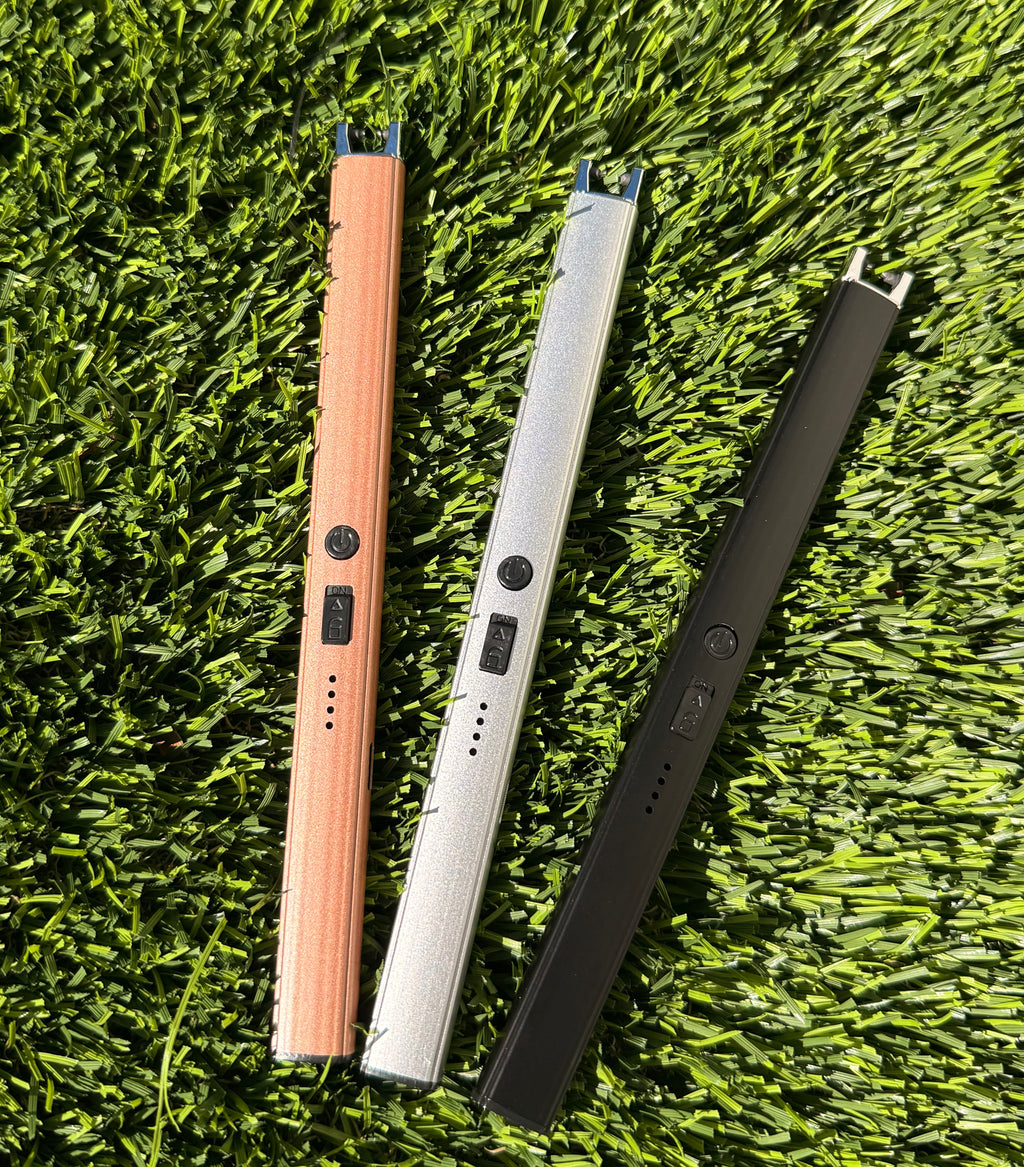 A picture of three electric lighters laying on a grass background. One is bronze, one is silver, and one is black.