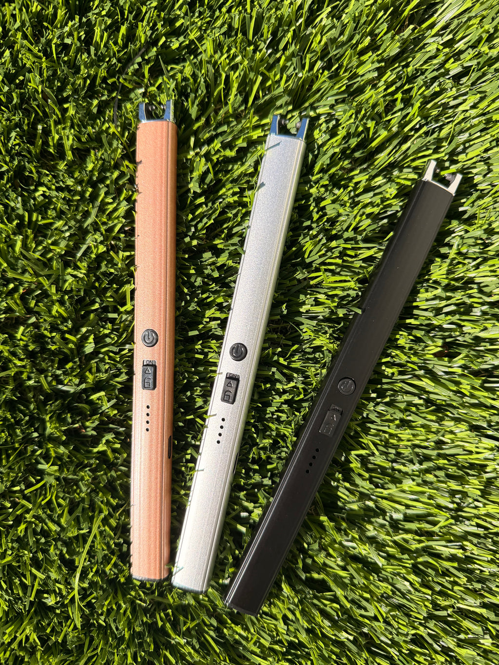 A picture of three electric lighters laying on a grass background. One is bronze, one is silver, and one is black.