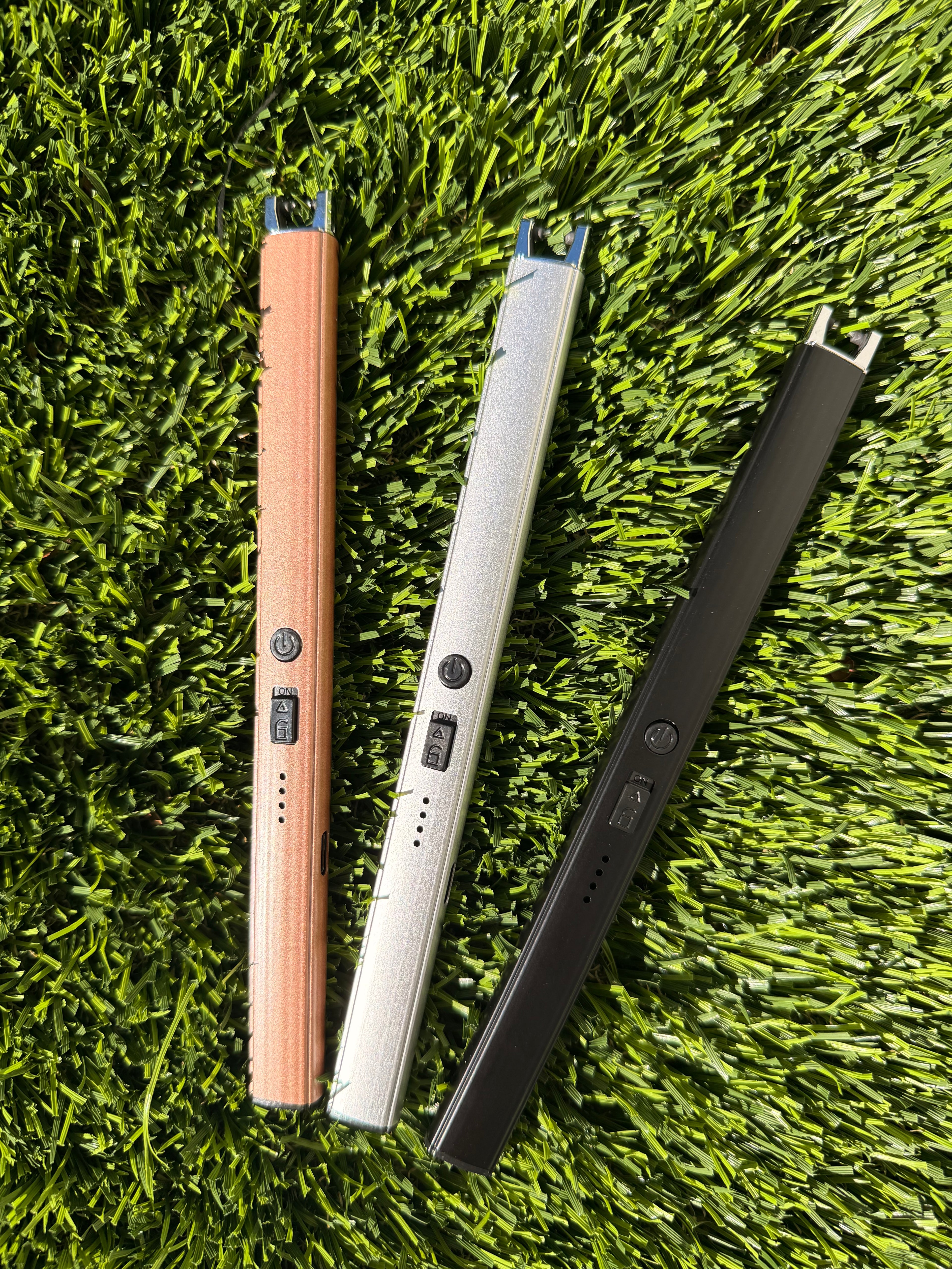 A picture of three electric lighters laying on a grass background. One is bronze, one is silver, and one is black.