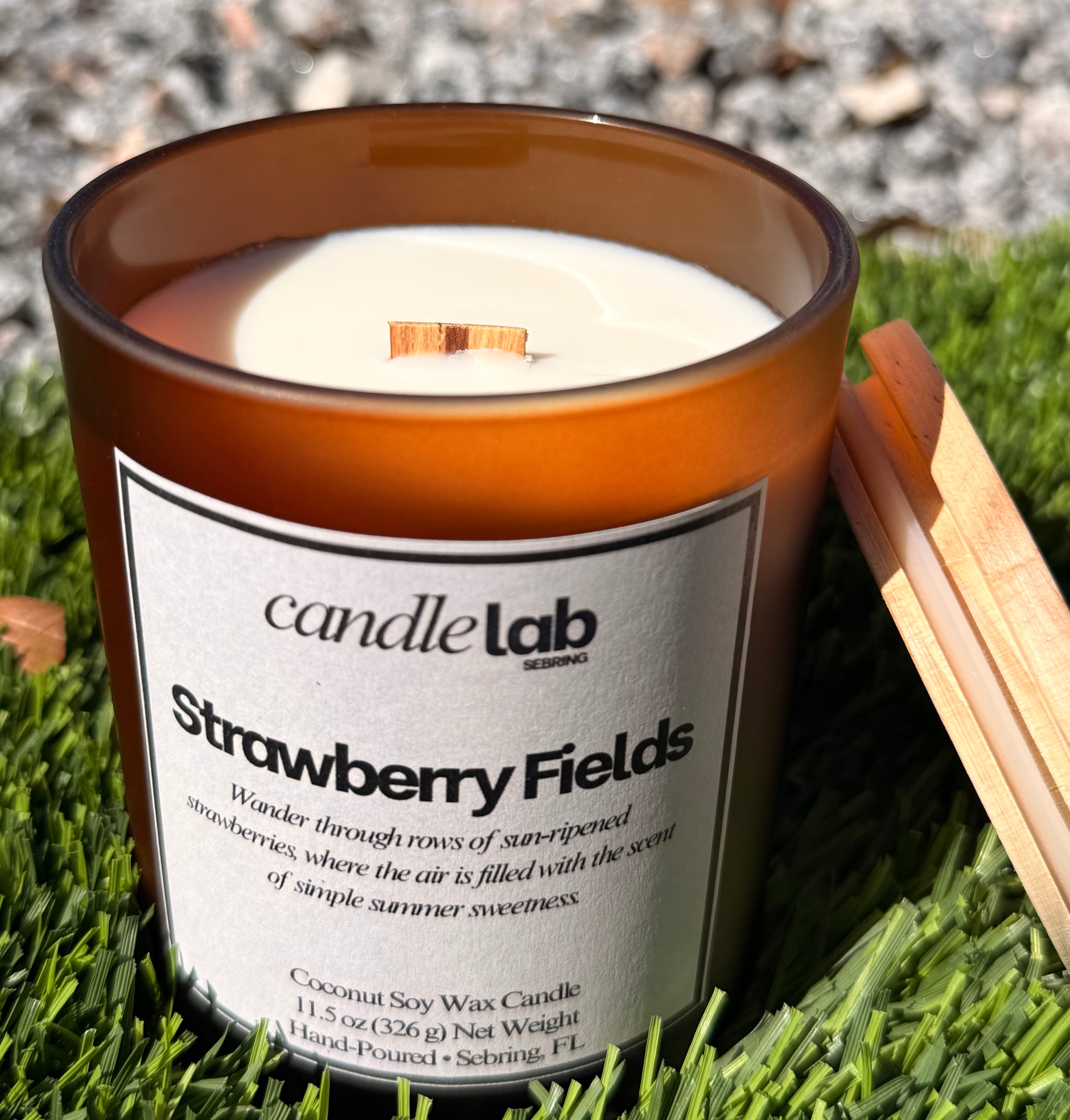 An amber glass candle with a label that says "Strawberry Fields" along with a short blurb reading "Wander through rows of sun-ripened strawberries, where the air is filled with the scent of simple summer sweetness."