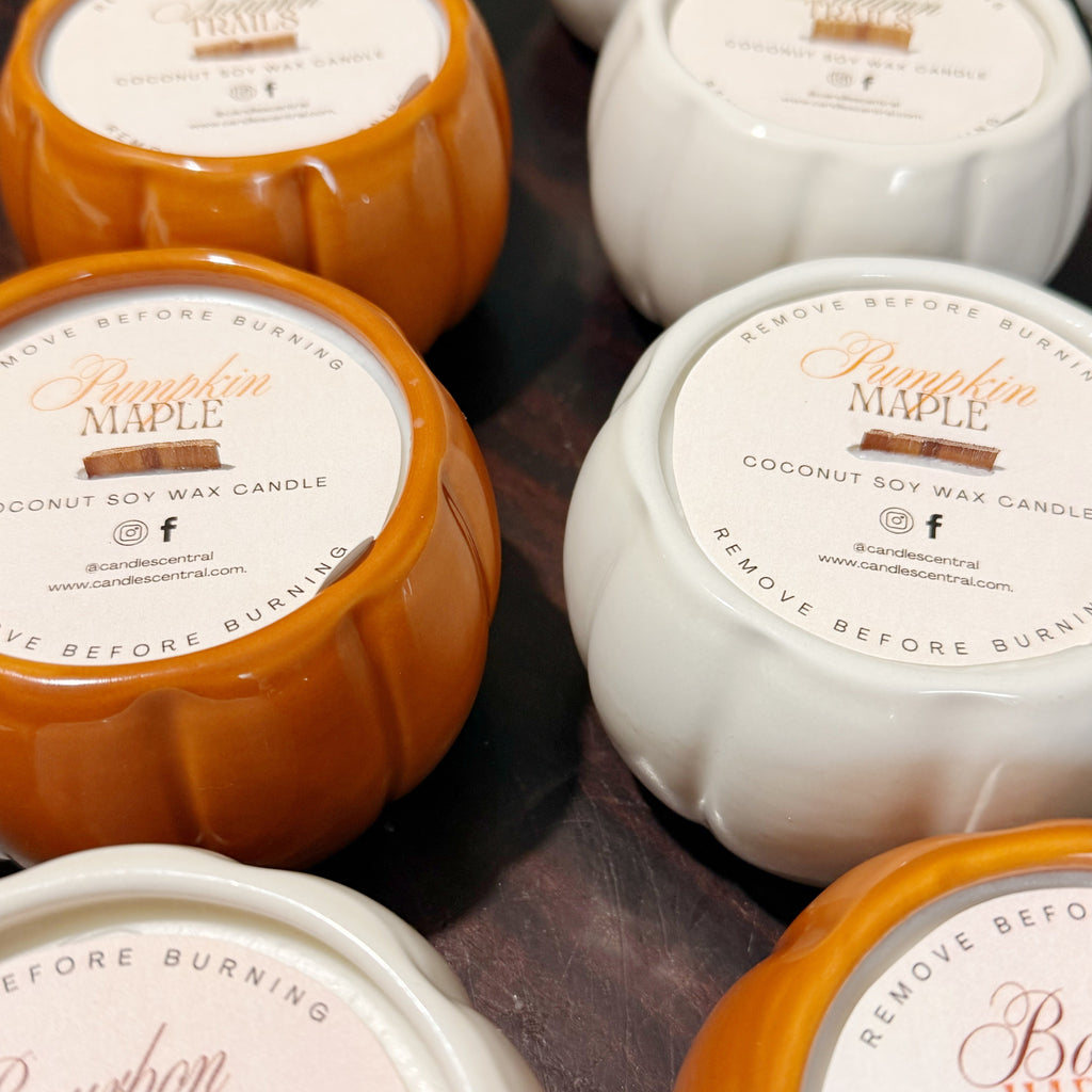 Several pumpkin-shaped coconut soy wax candles arranged on a dark surface. The visible candle bodies are orange and white, with round cream labels reading Pumpkin Maple and Bourbon Harvest.