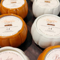 Several pumpkin-shaped coconut soy wax candles arranged on a dark surface. The visible candle bodies are orange and white, with round cream labels reading Pumpkin Maple and Bourbon Harvest.