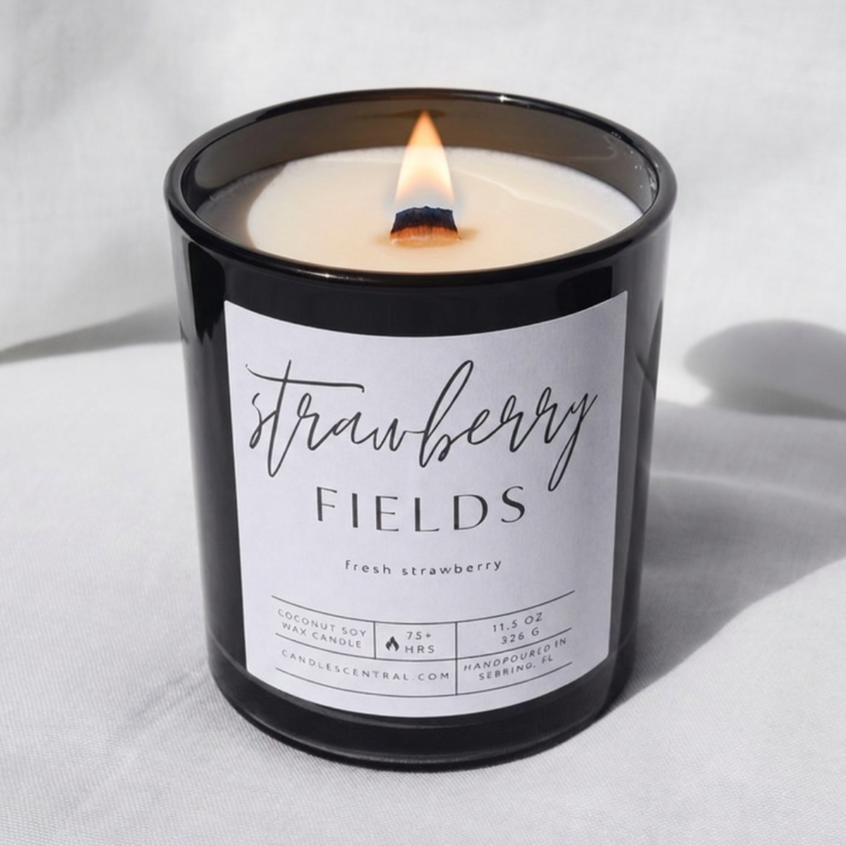 Picture of a candle burning with a label that says "Strawberry Fields" along with the fragrance note of fresh strawberry.