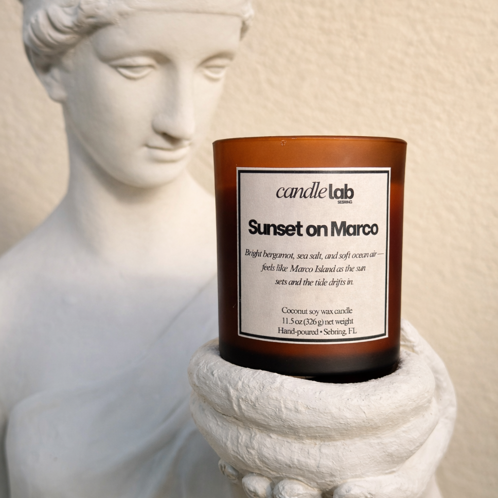 A picture of a statue holding an amber glass candle with a label that says "Sunset on Marco" along with a short blurb reading "Bright bergamot, sea salt, and soft ocean air-- feels like Marco Island as the Sun sets and the tide drifts in."