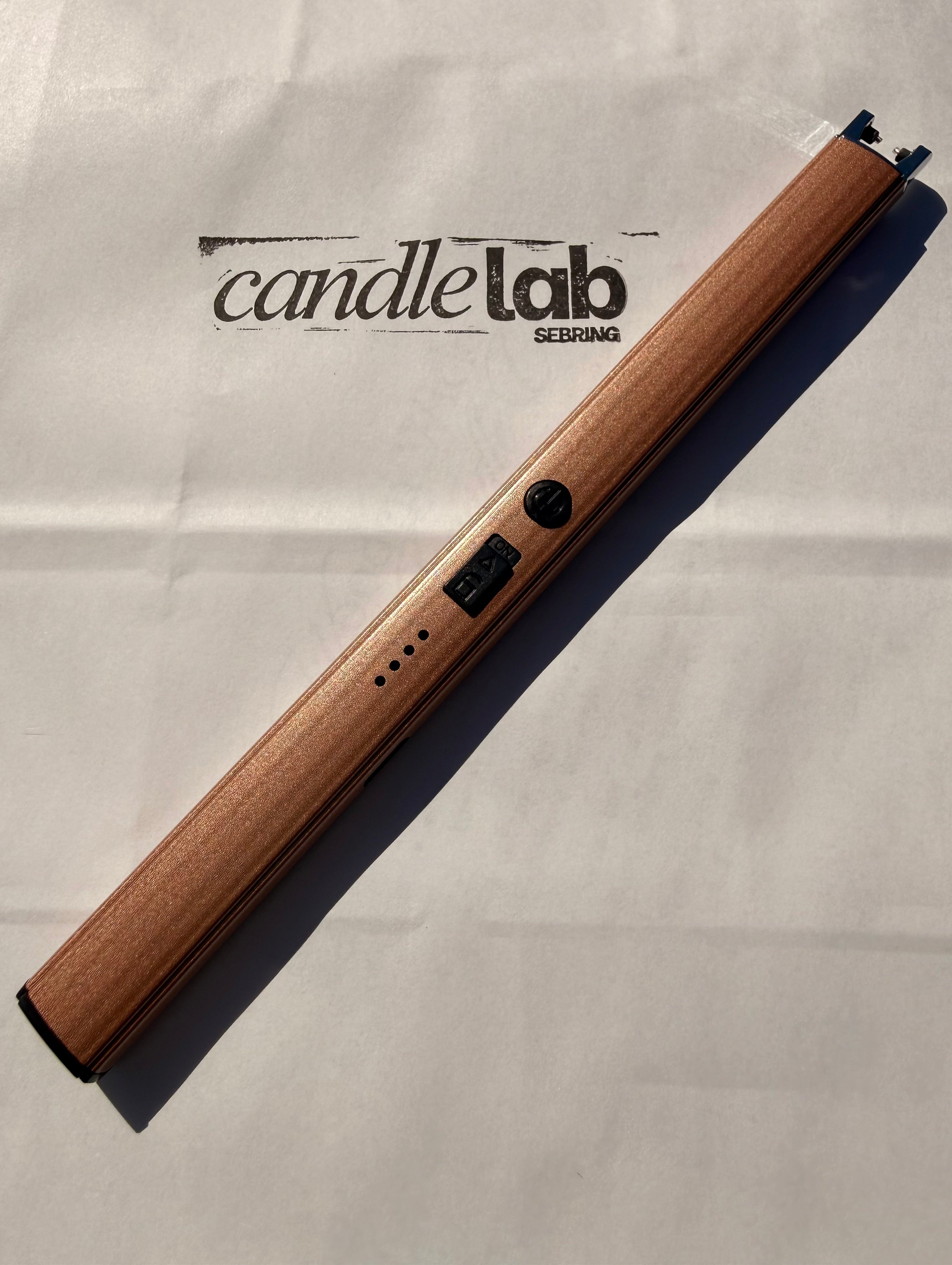 A picture of a bronze electric lighter laying on a white background with the words "Candle Lab Sebring" printed on the background.