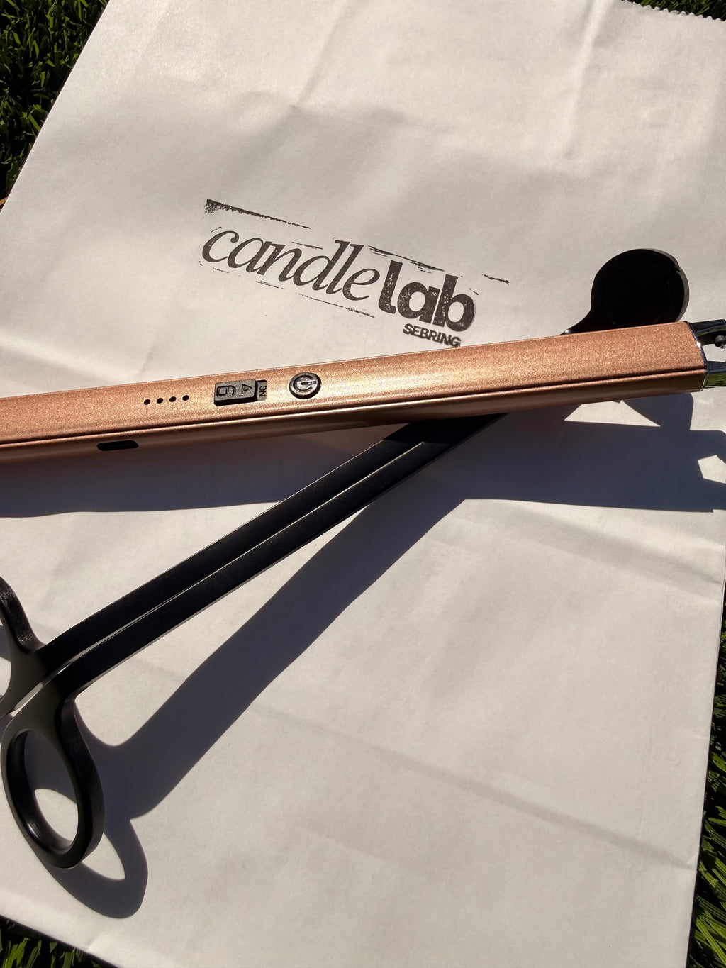 A picture of a bronze electric lighter and black wick trimmer sitting on a white bag with the words "Candle Lab Sebring" printed on the background.